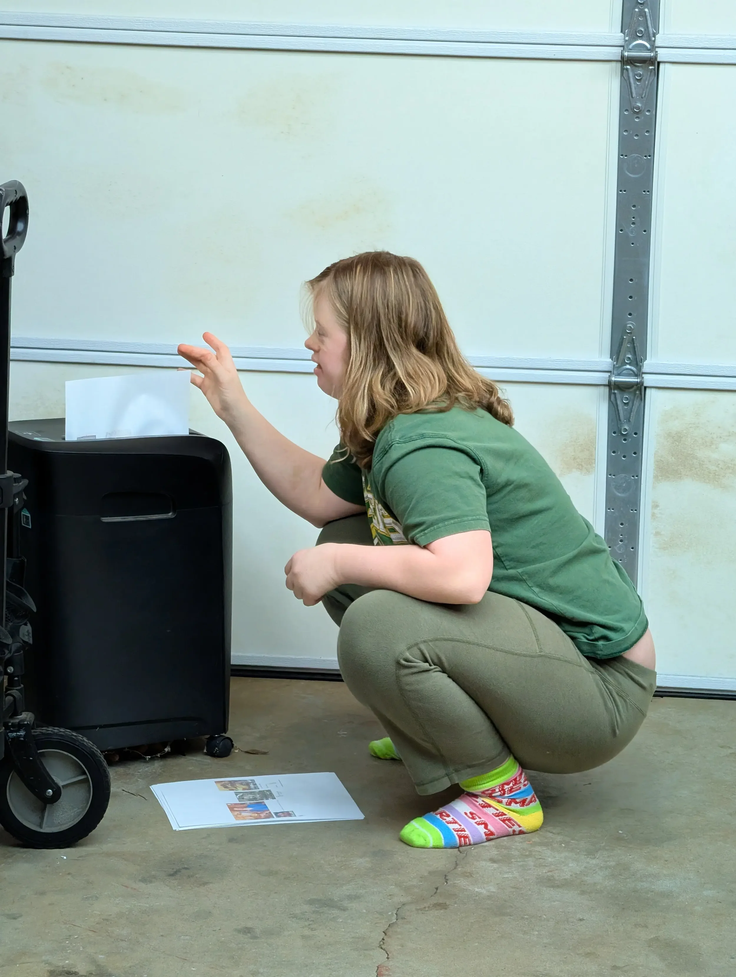A woman crouching in a garage near a black shredder, with a sheet of photo prints on the floor in front of her.