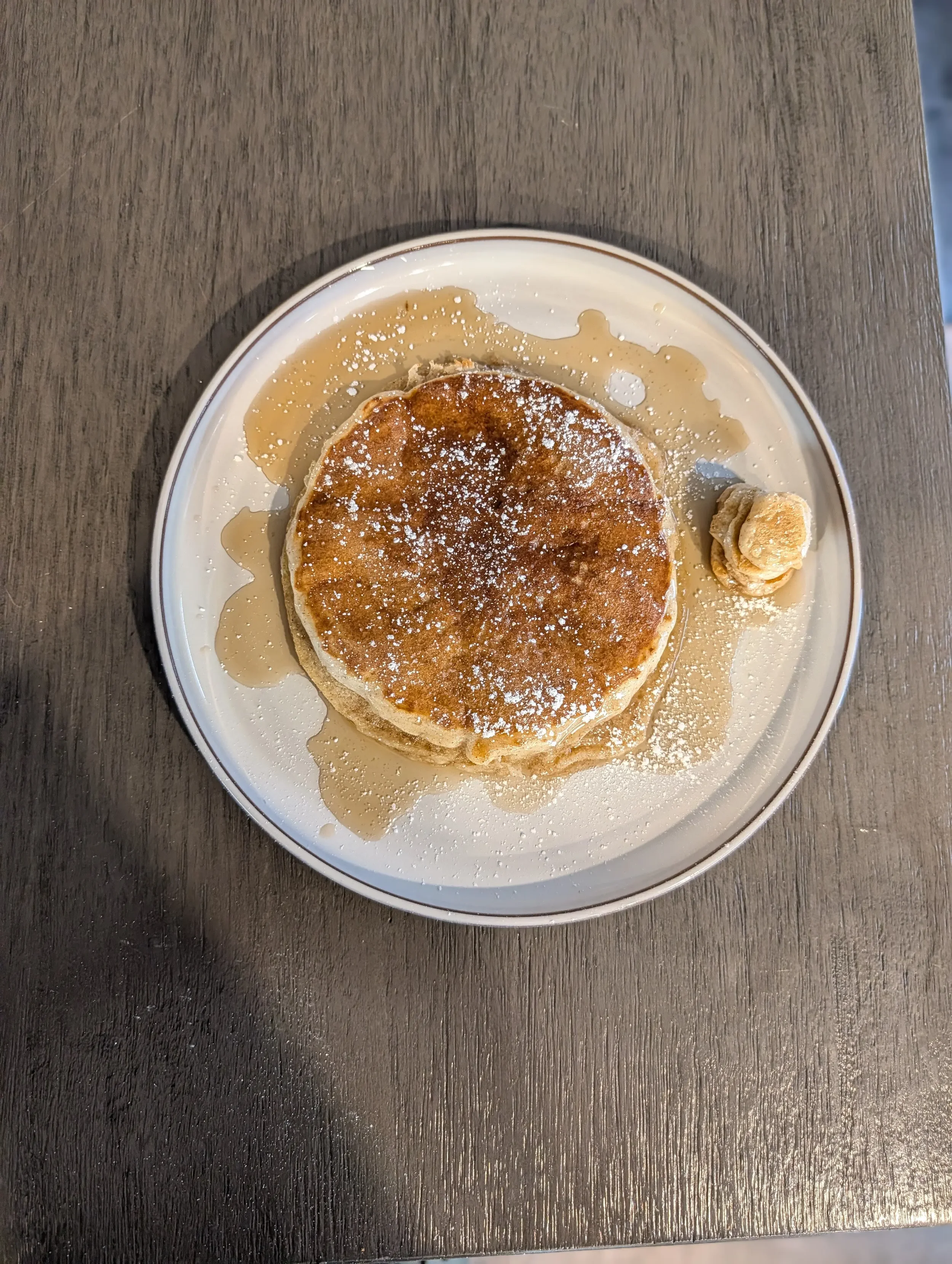 A plate of pancakes topped with powdered sugar, with syrup spilled around the pancake, placed on a dark wooden surface.