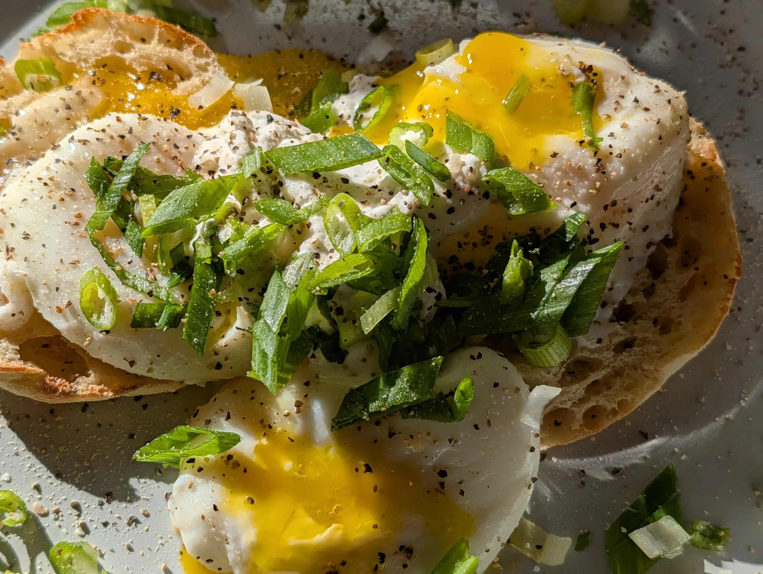 Close-up of an open-faced breakfast sandwich with poached eggs, chopped green onions, black pepper, and seasoning on toasted bread.