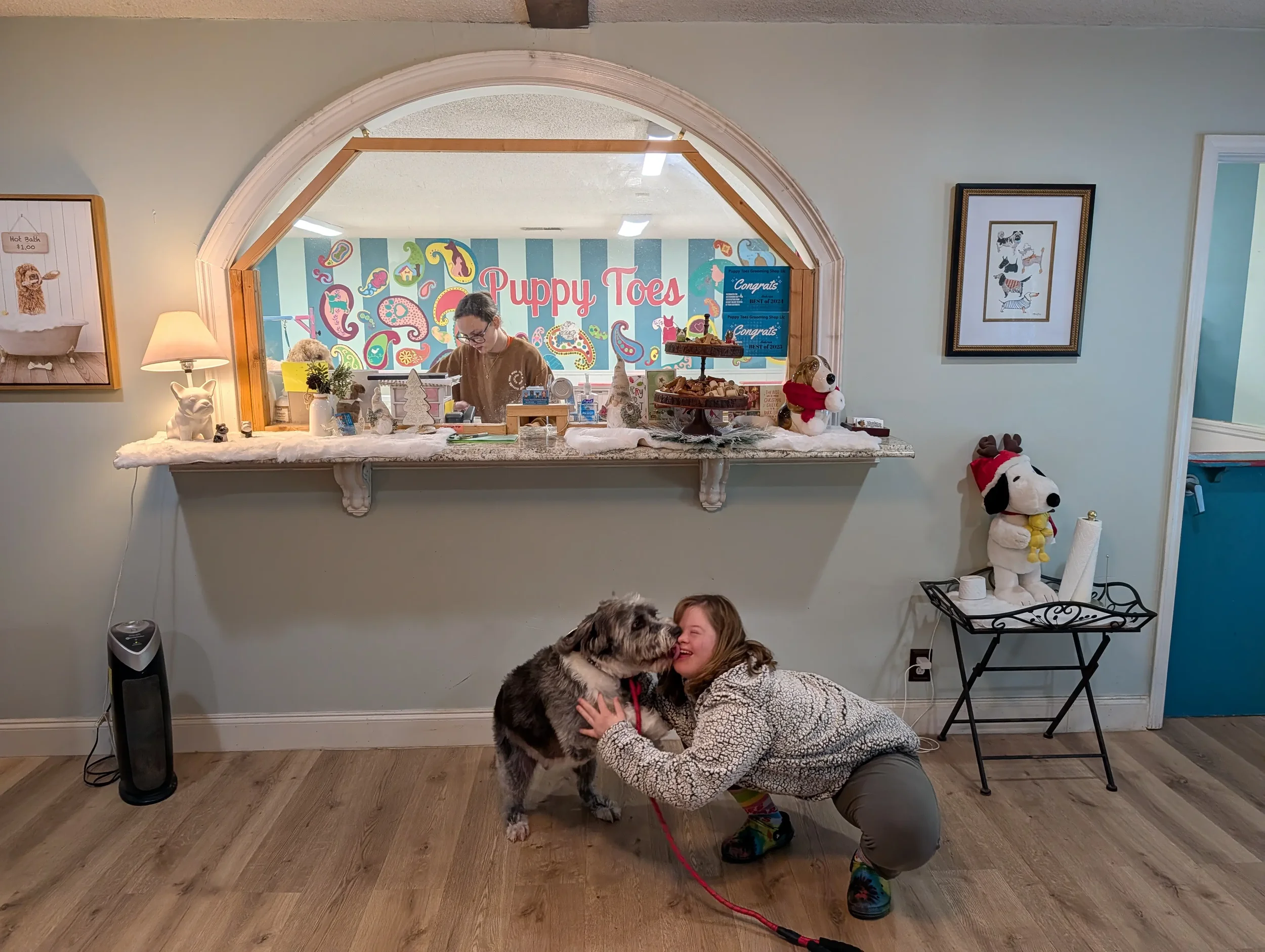 A girl hugging and kissing a large, fluffy dog in a room decorated with Christmas items. In the background, there is a pet grooming salon window with a sign that reads 'Puppy Toes', and the room has Christmas decorations and Peanuts characters like S