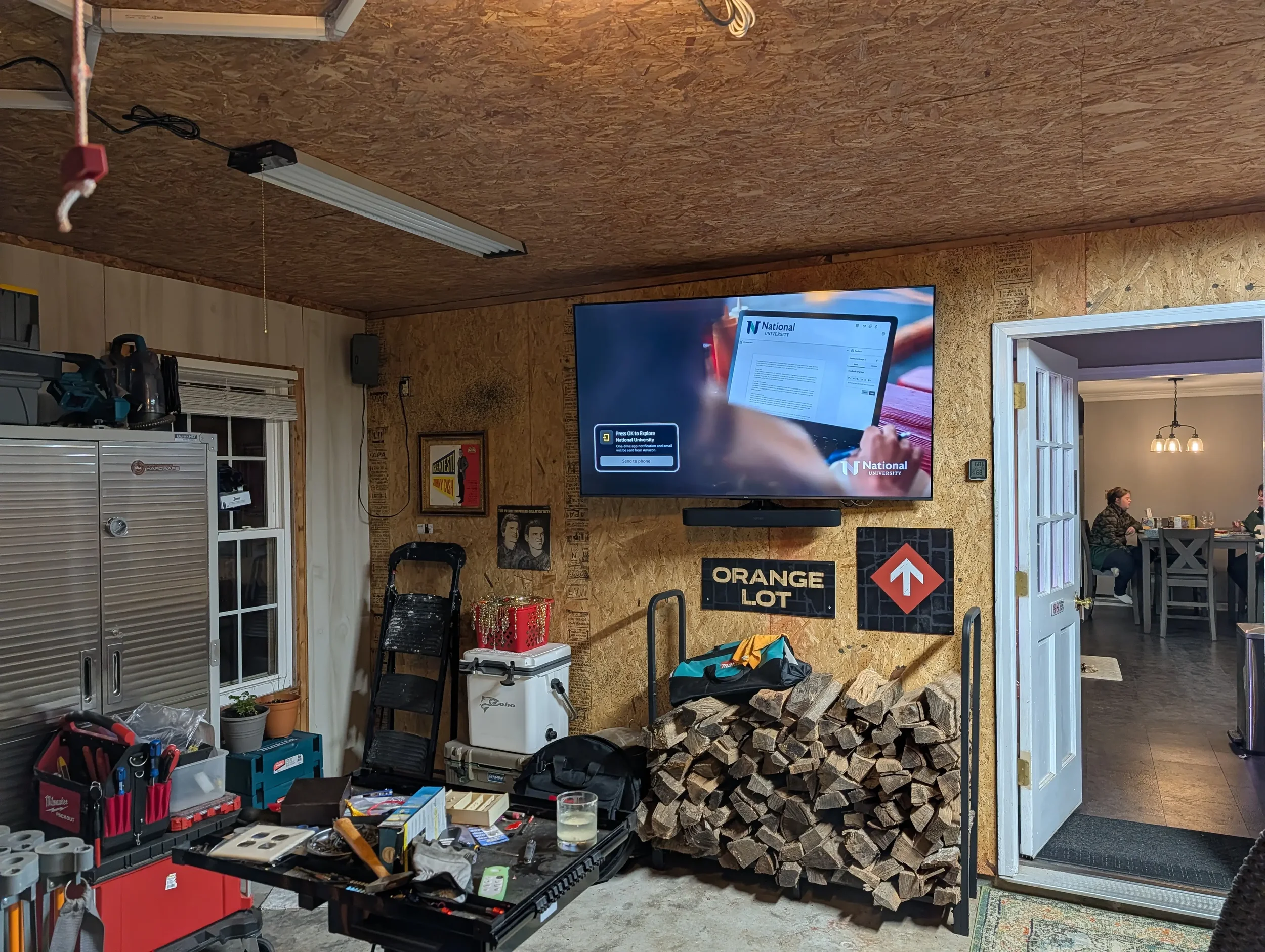 Indoor workshop or garage with a large TV mounted on a wood wall, displaying a tablet with a university logo. Below the TV, firewood is stacked, and there are signs reading 'ORANGE LOT' and an arrow pointing upward. The room contains tools, storage c
