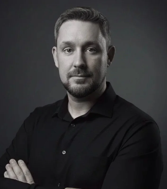 A black-and-white portrait of a man with short hair and a beard, wearing a black button-up shirt with arms crossed, looking at the camera with a neutral expression.