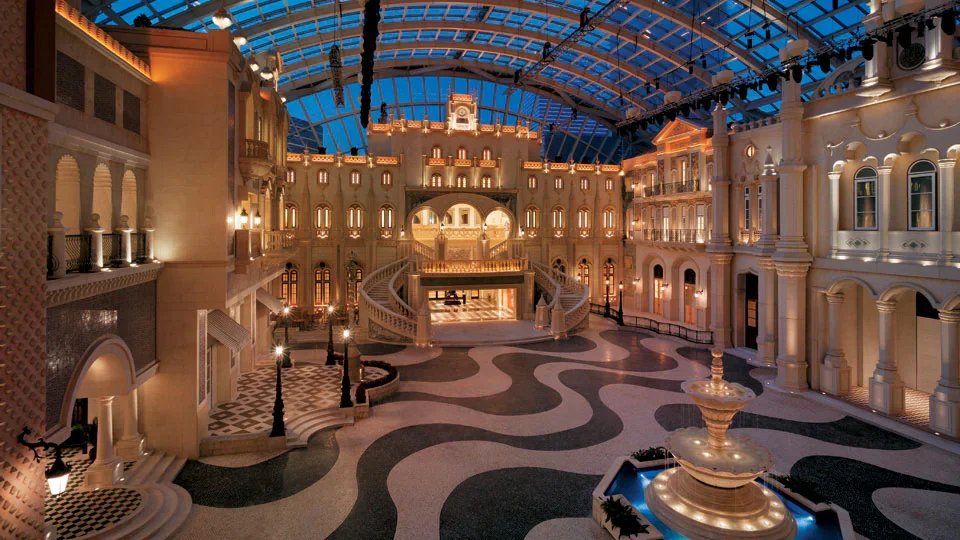 Interior view of a large, elegant hotel lobby with ornate architecture, grand staircase, and a decorative fountain, illuminated with warm lighting under a glass-domed ceiling.