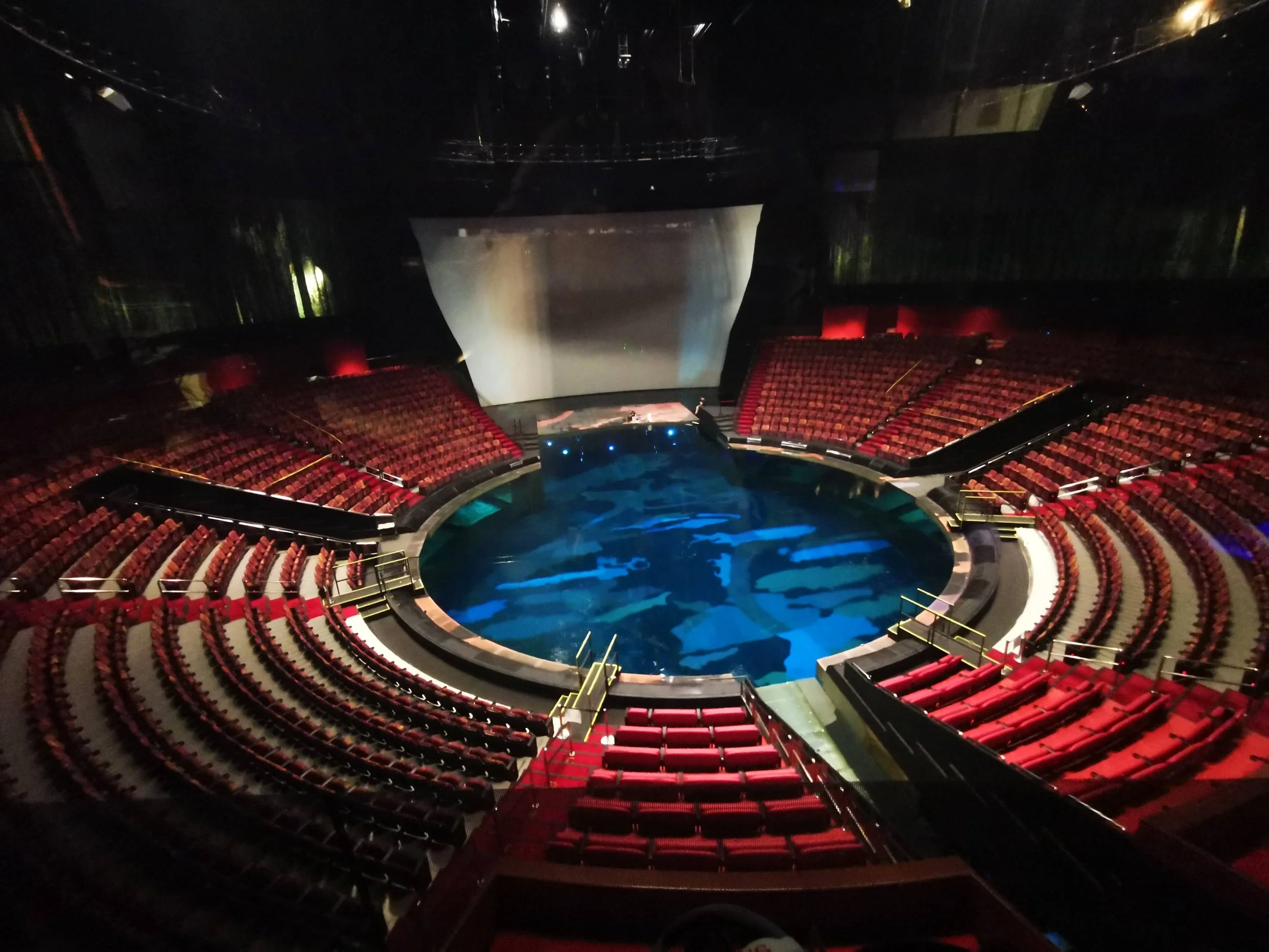 An empty theater with red seats arranged in a semi-circle around a stage with a blue water feature, dark lighting, and a large screen behind the stage.