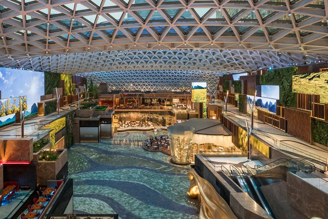Interior of a modern, multi-level shopping mall with a high, geometric ceiling, large digital screens on the walls, and a central area featuring a water feature and escalators.