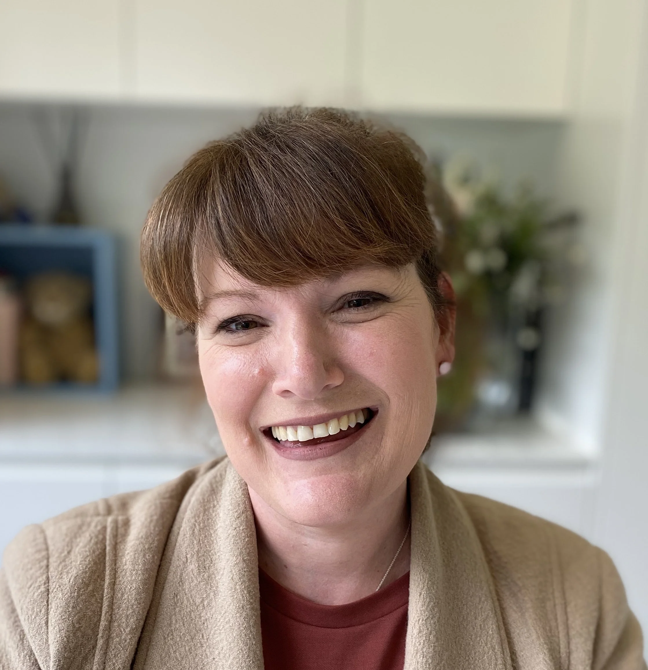 Close-up of a smiling woman with short brown hair, wearing a beige blazer and red top, in a well-lit room with a blurred background.