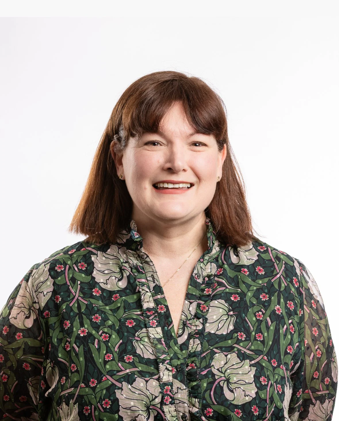 A woman with brown hair and a smile, wearing a floral patterned blouse, standing against a plain white background.
