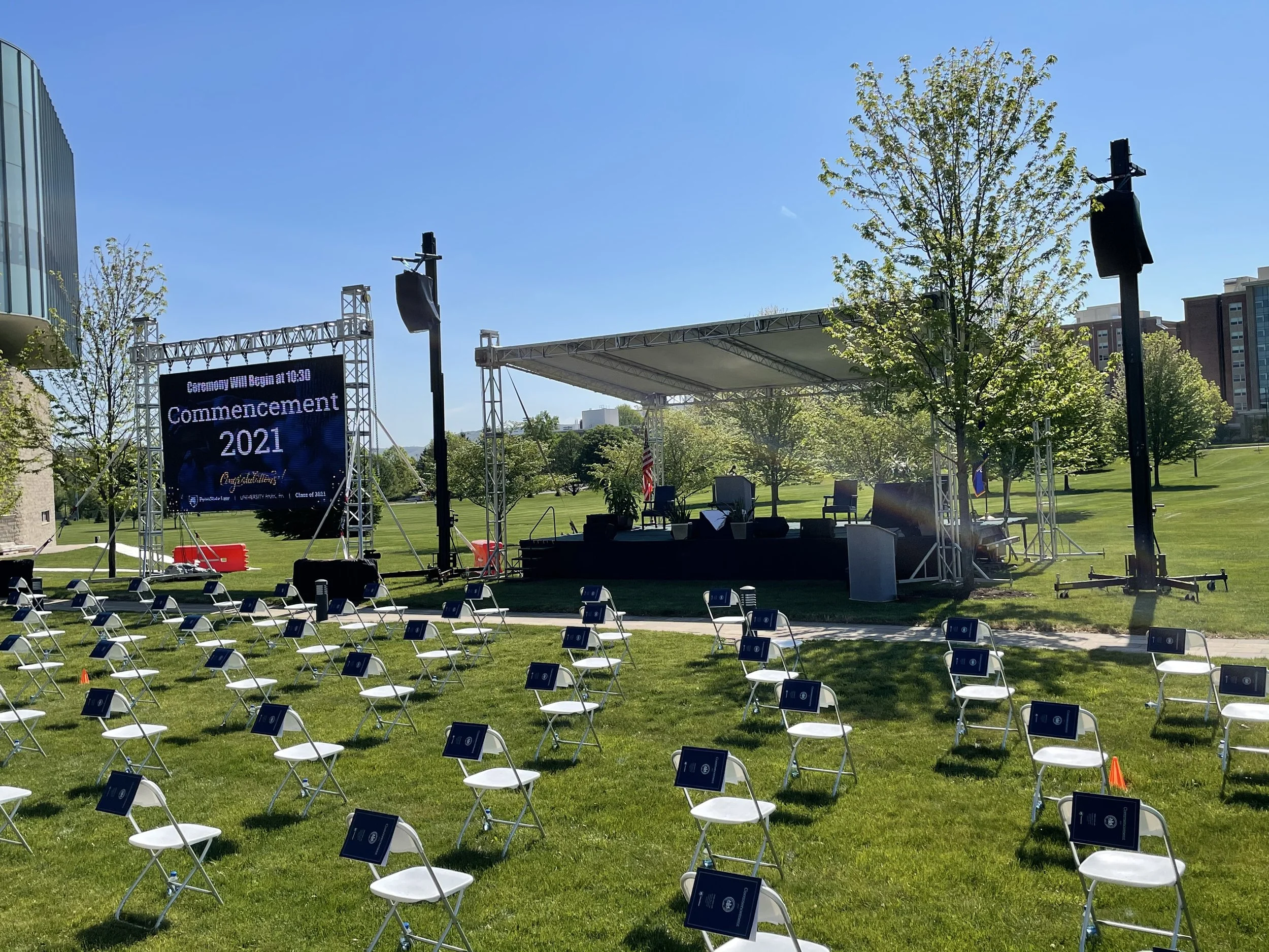 An outdoor stage set up for a graduation ceremony with chairs arranged in front and a large digital screen displaying 'Commencement 2021' on a bright, sunny day with a clear blue sky.