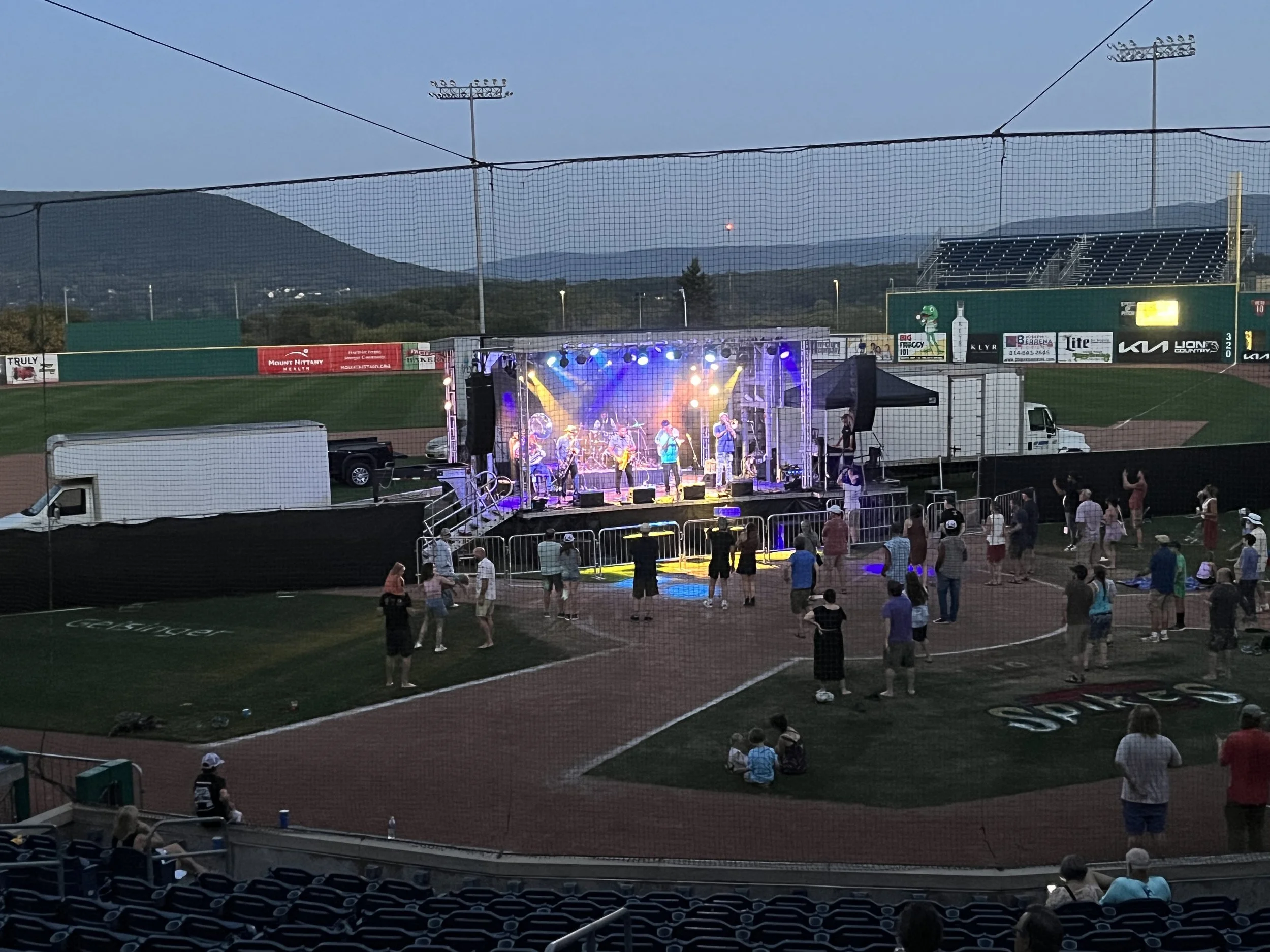 People watching a live band on a stage at a baseball stadium in the evening, with the field visible in the background and a mountain range beyond.
