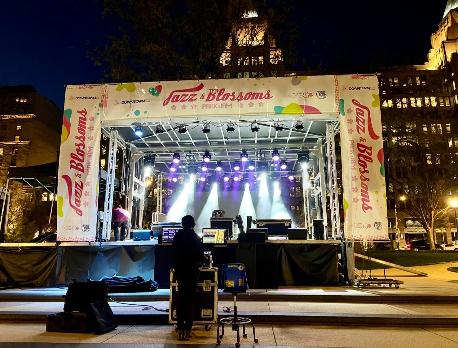 Empty outdoor stage with lighting and sound equipment at night, set up for a music event called Jazz & Blossoms Park Jam in downtown.