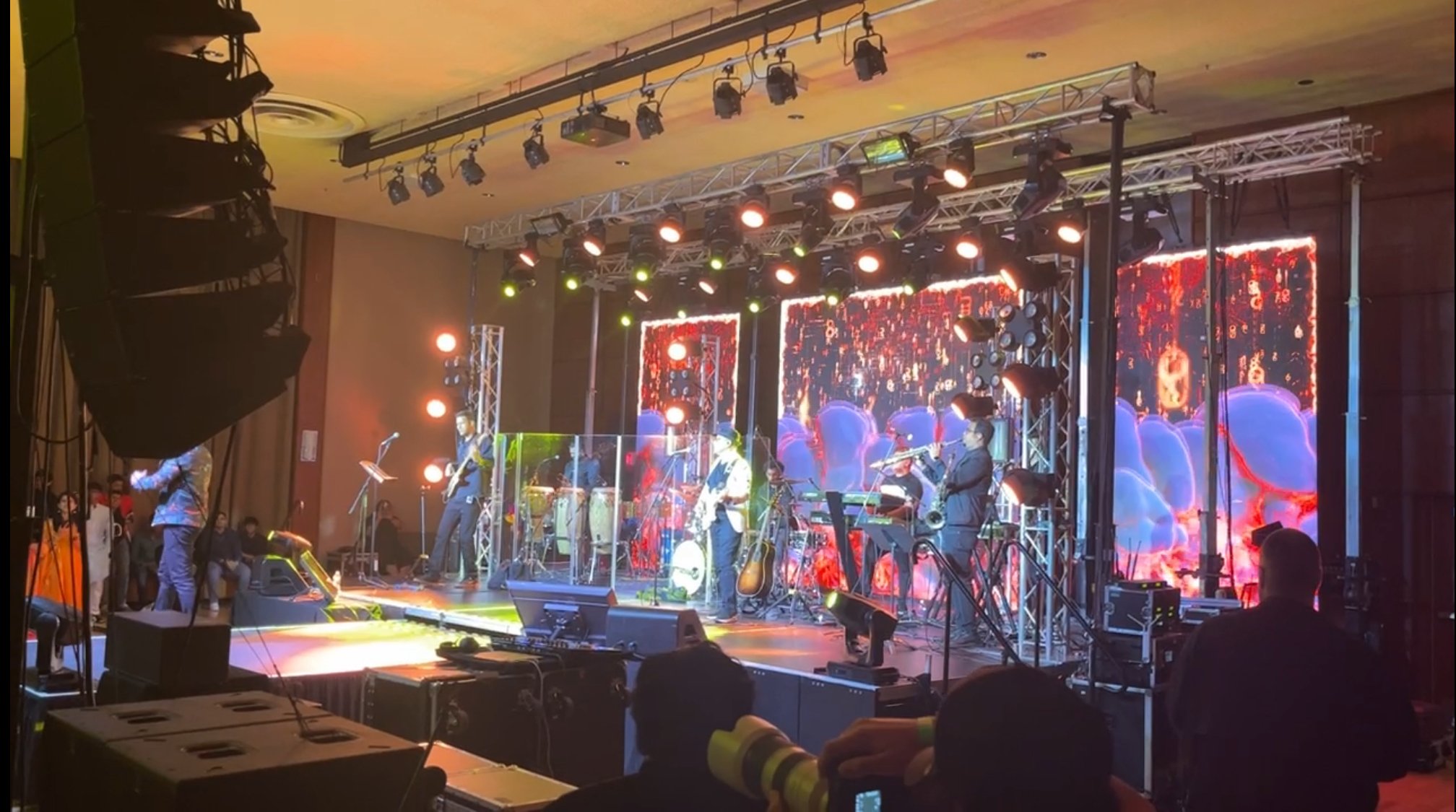 A live band performing on a stage with colorful electronic screens and stage lighting, with an audience and crew members watching.