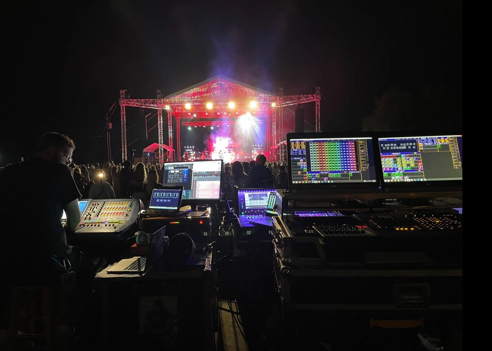 A night concert scene with a stage lit up with lights and smoke. In the foreground, a sound engineer operates multiple sound and lighting control panels and computers.