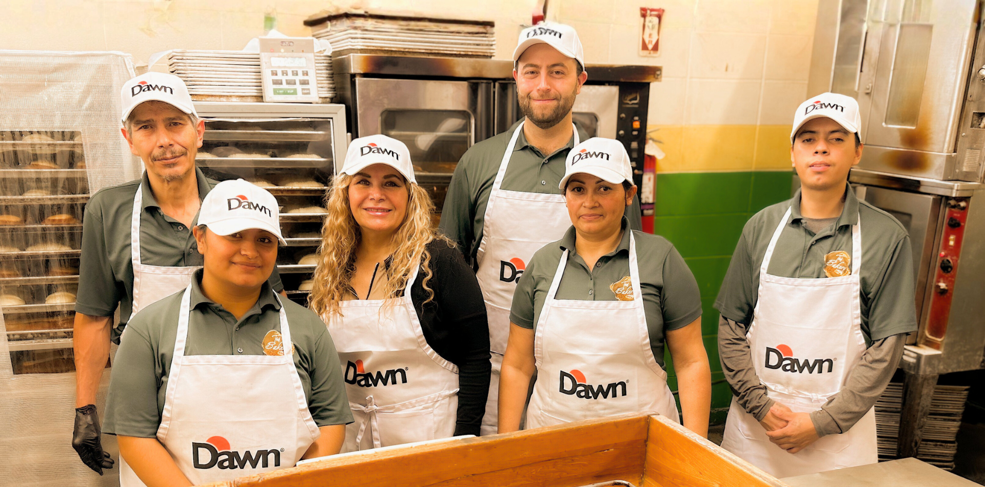 Group of bakery employees standing together in a bakery kitchen, wearing Dawn and bakery aprons and hats, smiling for the camera.