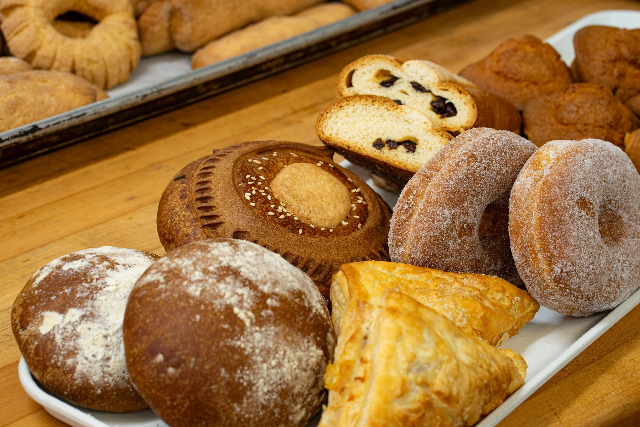 Assorted baked goods on a white platter, including donuts, Danish pastries, and cookies, with additional baked goods on a tray in the background.
