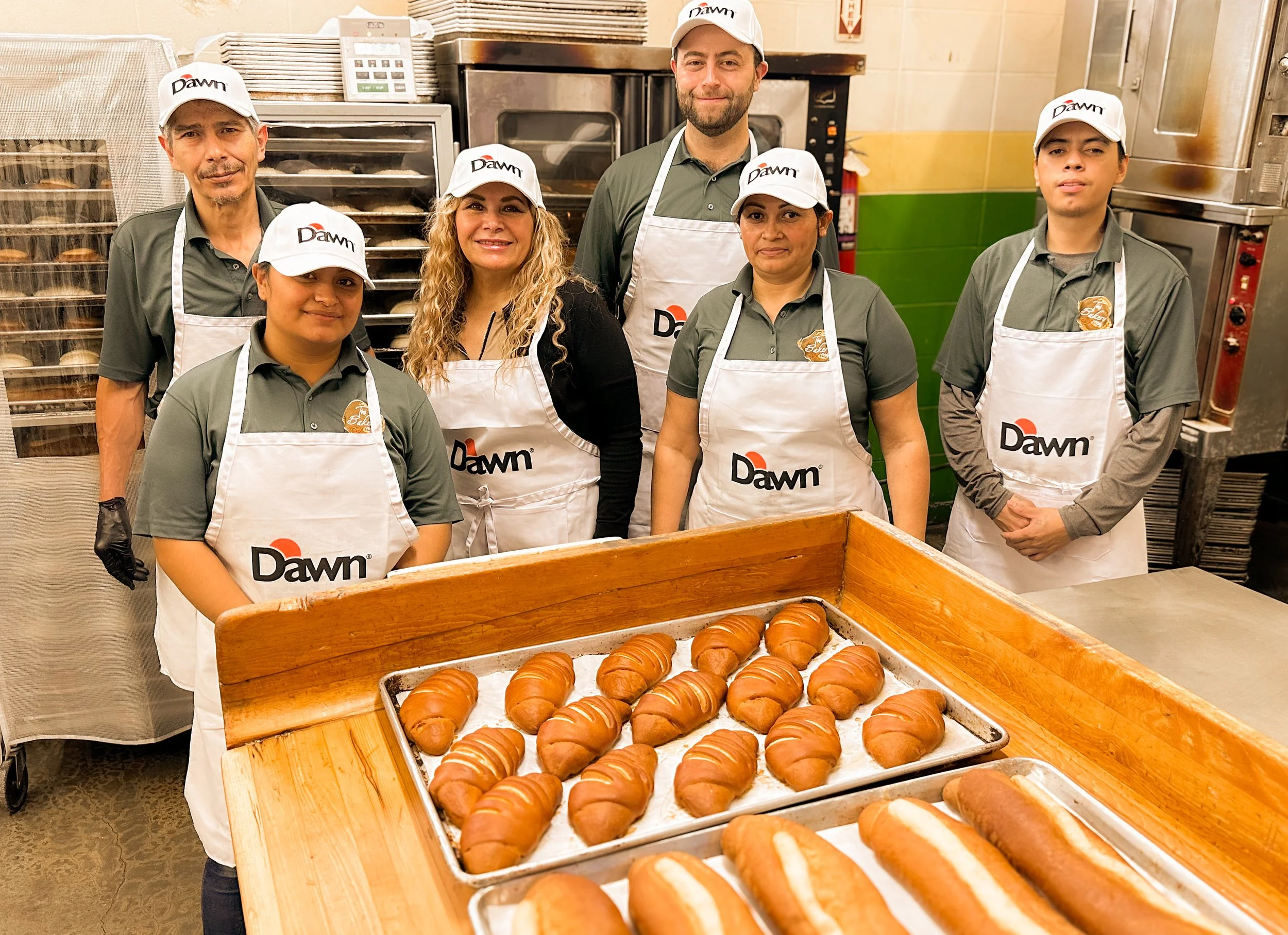 Group of six bakery workers in uniform, standing behind a table of freshly baked bread and croissants in a commercial bakery kitchen.