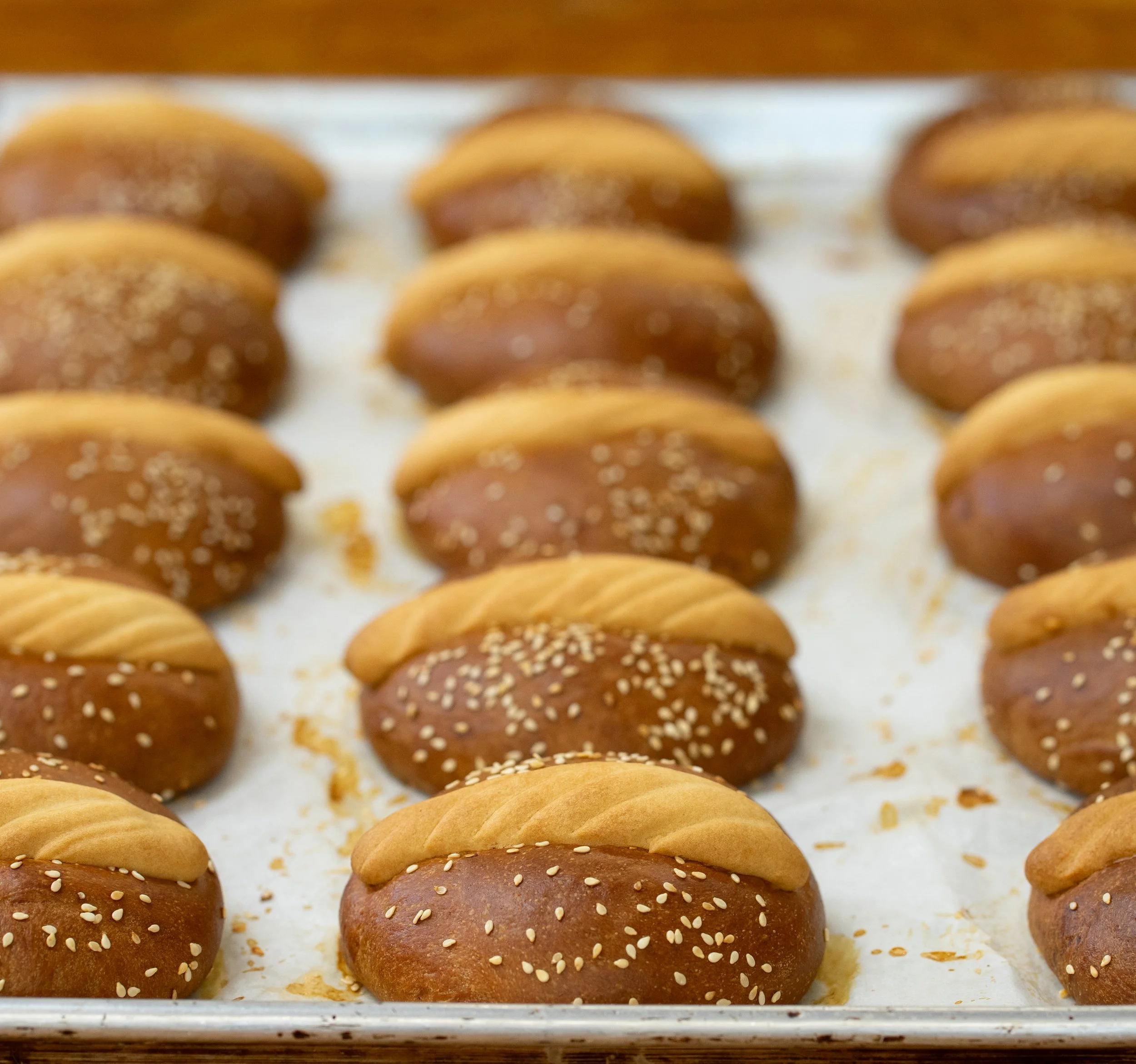 Baked bread rolls topped with sesame seeds arranged on a baking tray.