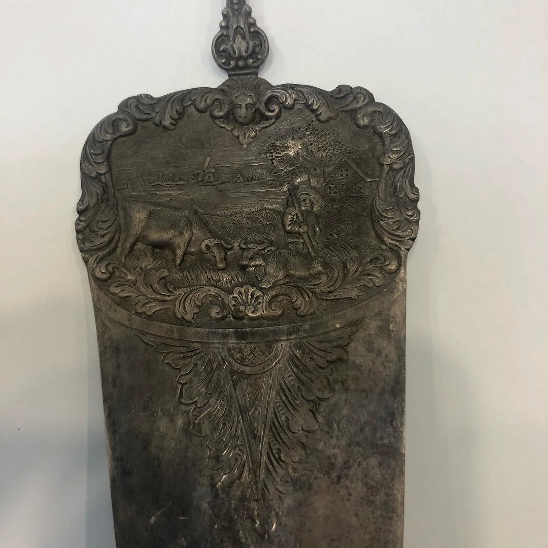 Decorative silver cake server with an engraved outdoor scene of a person farming, cows, a house, and trees.