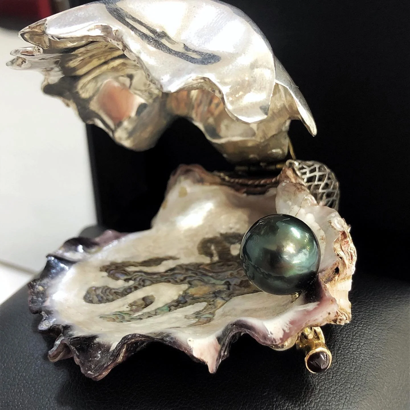 A large oyster shell with a dark pearl resting in it and jewelry items, all placed on a black surface.