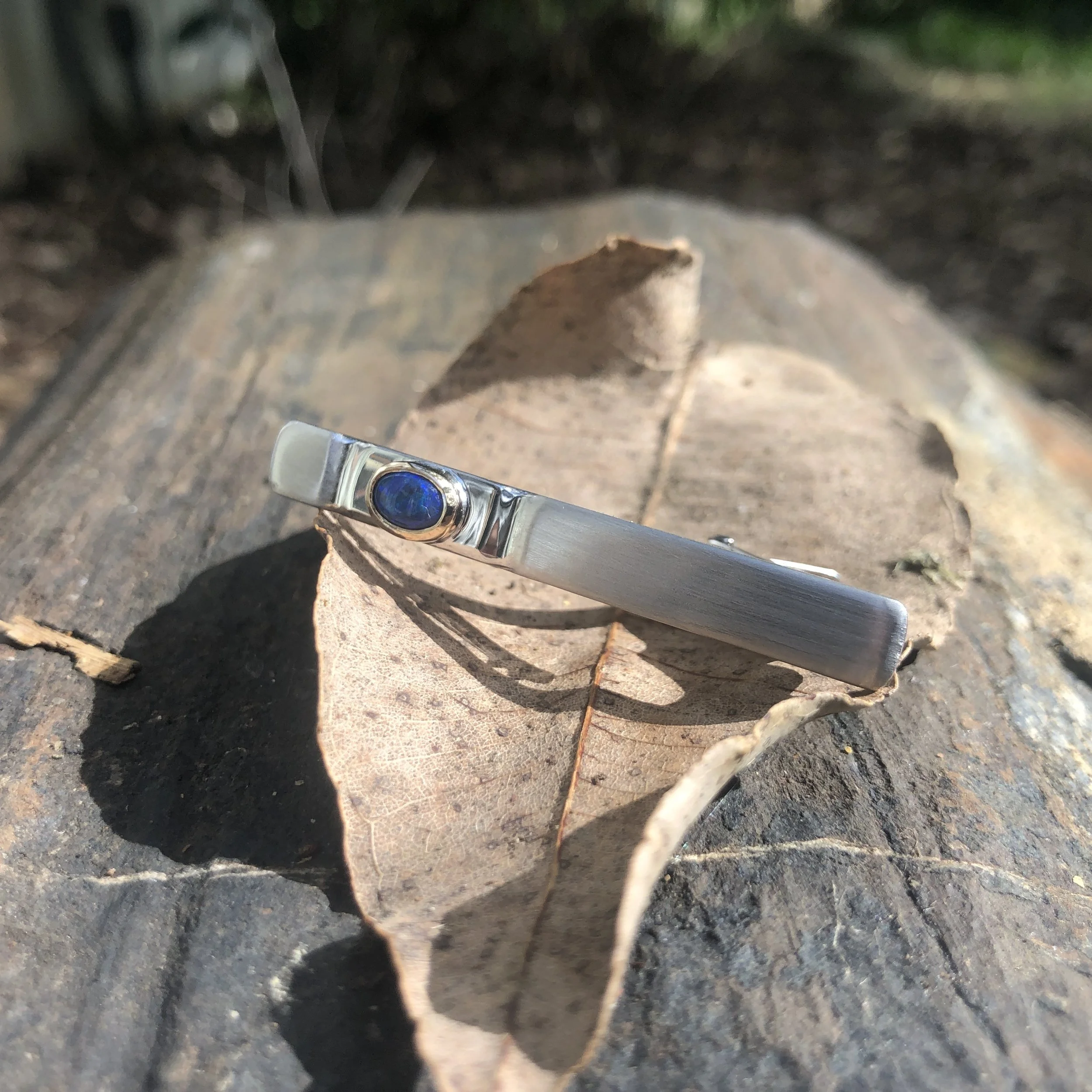 Silver tie pin with a blue gemstone, placed on a rough surface covered with a dried leaf and small rocks.