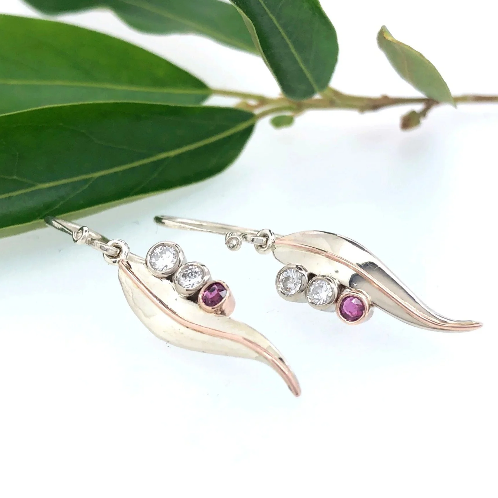 Silver earrings with pink and clear gemstones, placed on a light surface near green leaves.