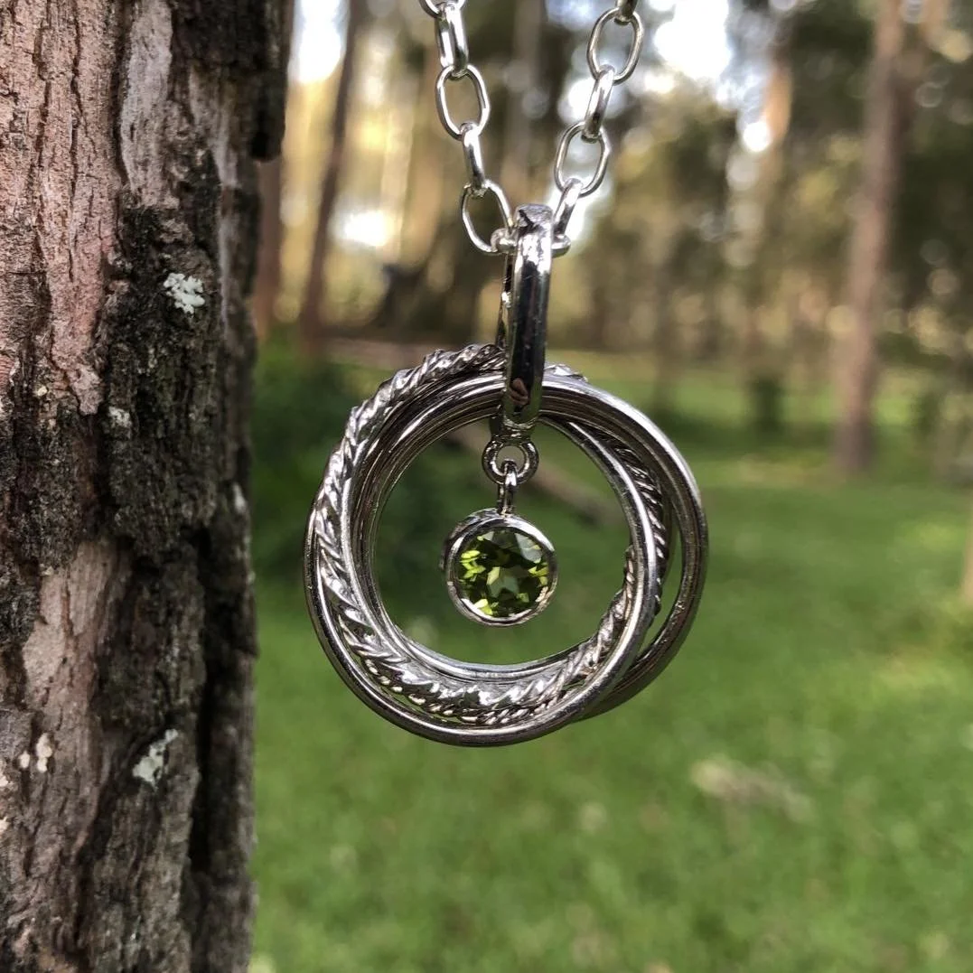 A close-up photo of a white gold necklace with concentric circular pendants and a green gemstone, hanging from a tree branch in a forest setting.