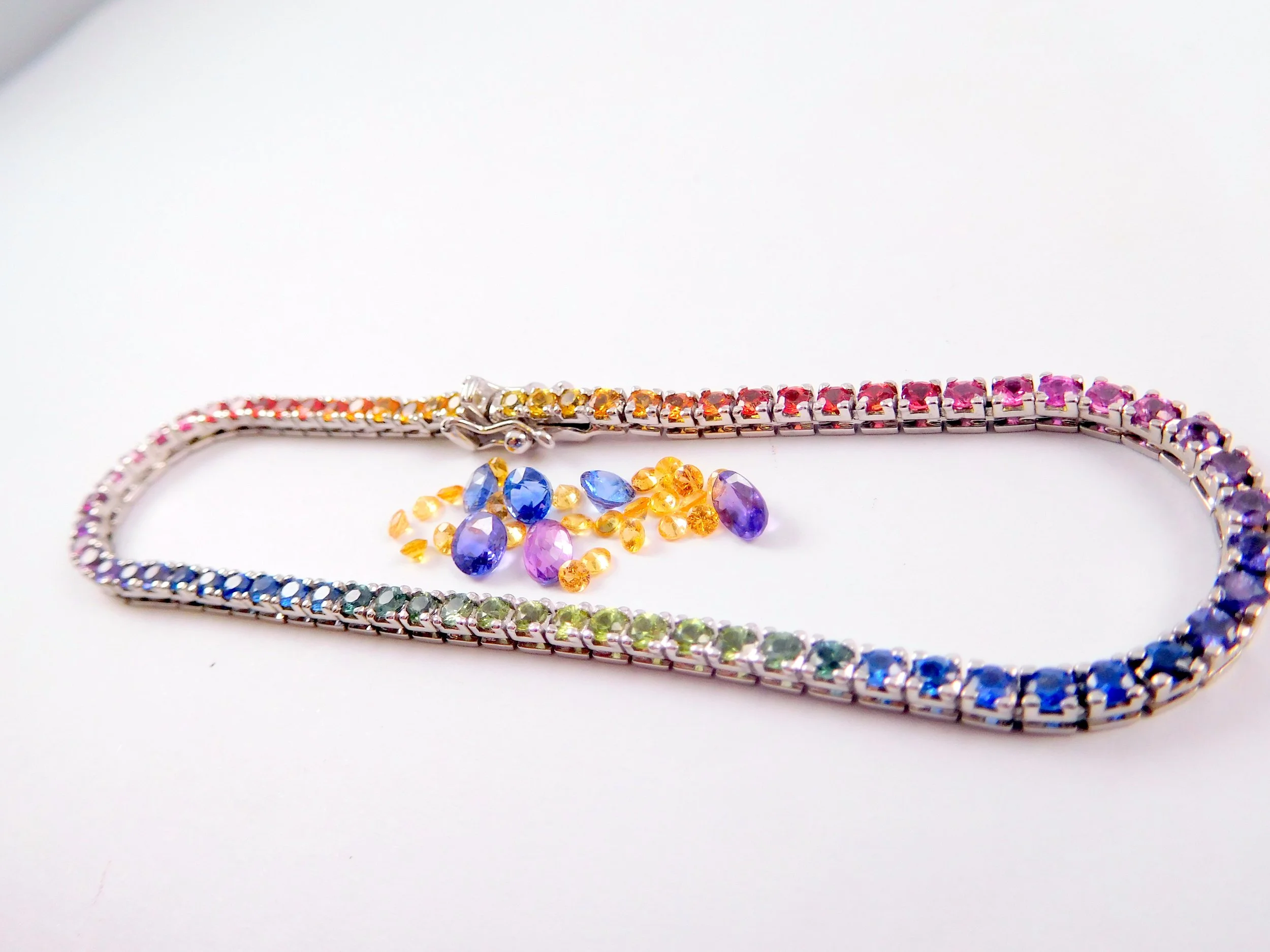 Colorful rainbow-colored jewelry: a bracelet with blue and purple stones, a bracelet with pink and purple stones, and a necklace with yellow and orange stones, all placed on a white surface.