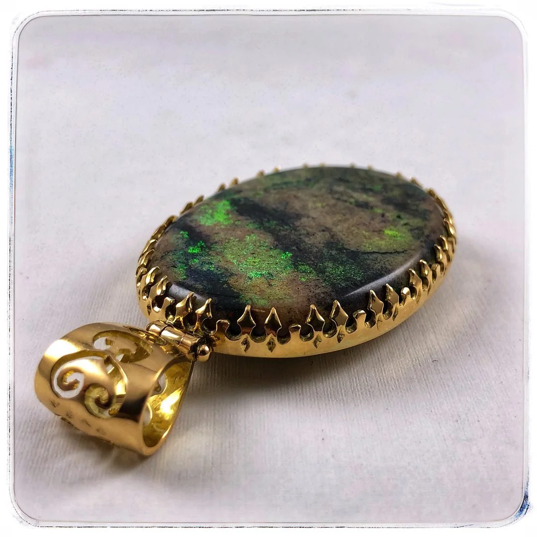 Gold pendant with an oval-shaped iridescent stone that has green and brown hues, set in a decorative gold frame with a scroll design on the bail.
