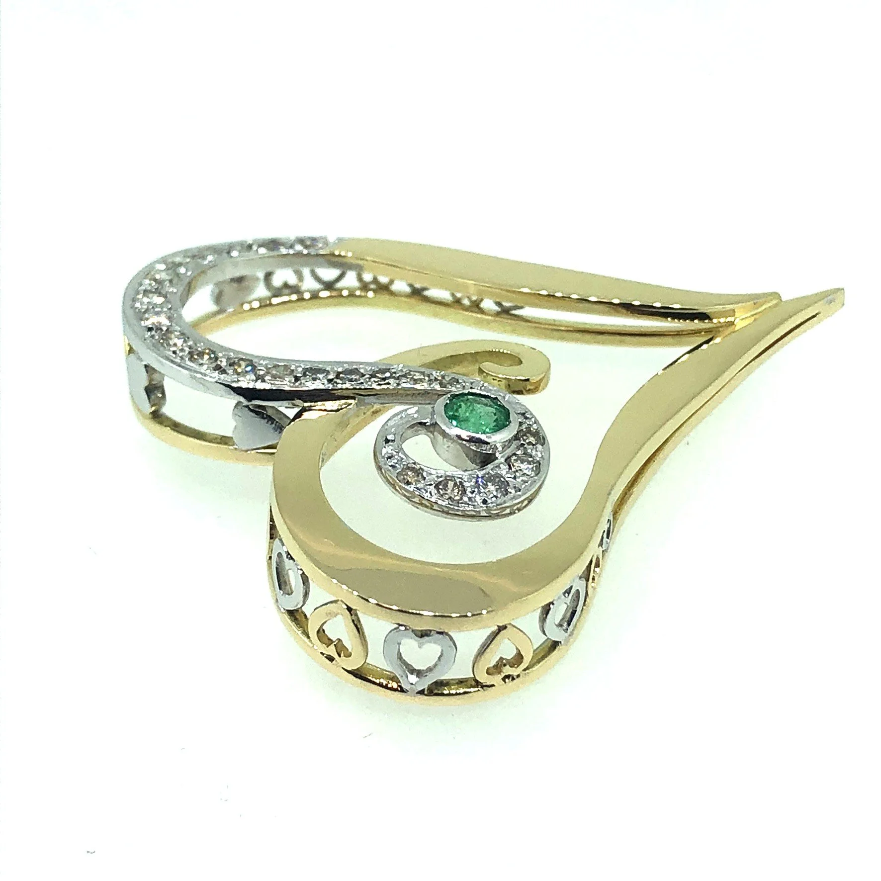 Close-up of a gold and white gold heart pendant - with intricate open-work design, a row of small diamonds, and a central green emerald gemstone, placed on a white surface.