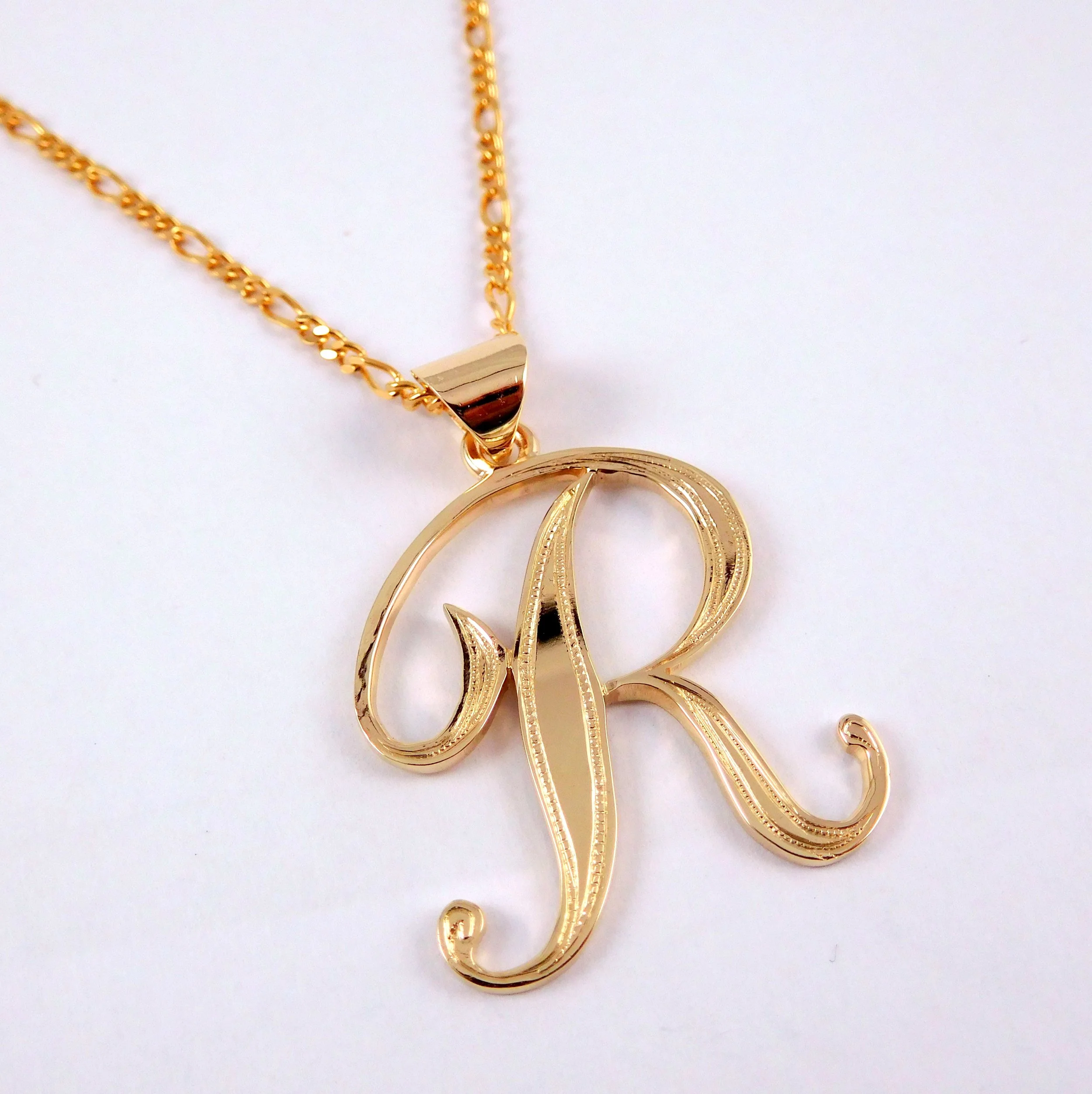 Close-up view of a gold necklace with a pendant in the shape of the letter 'R' in cursive script. 