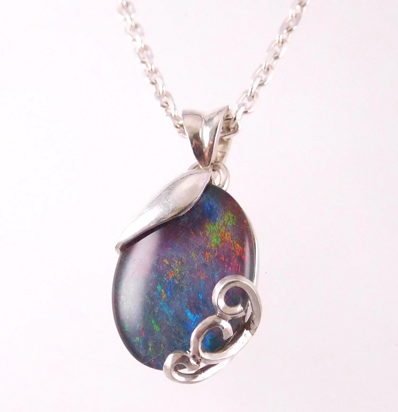 A silver necklace with a colorful, oval gemstone pendant featuring a swirling rainbow-like pattern, accented with silver wire wrapping.