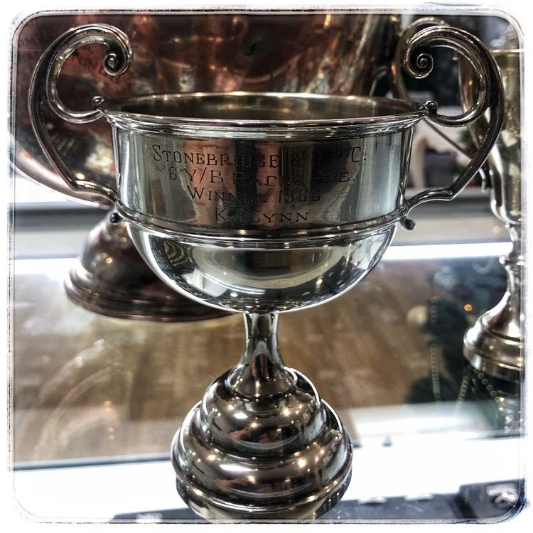 A silver trophy cup with engraved text, placed on a reflective surface, having been polished.