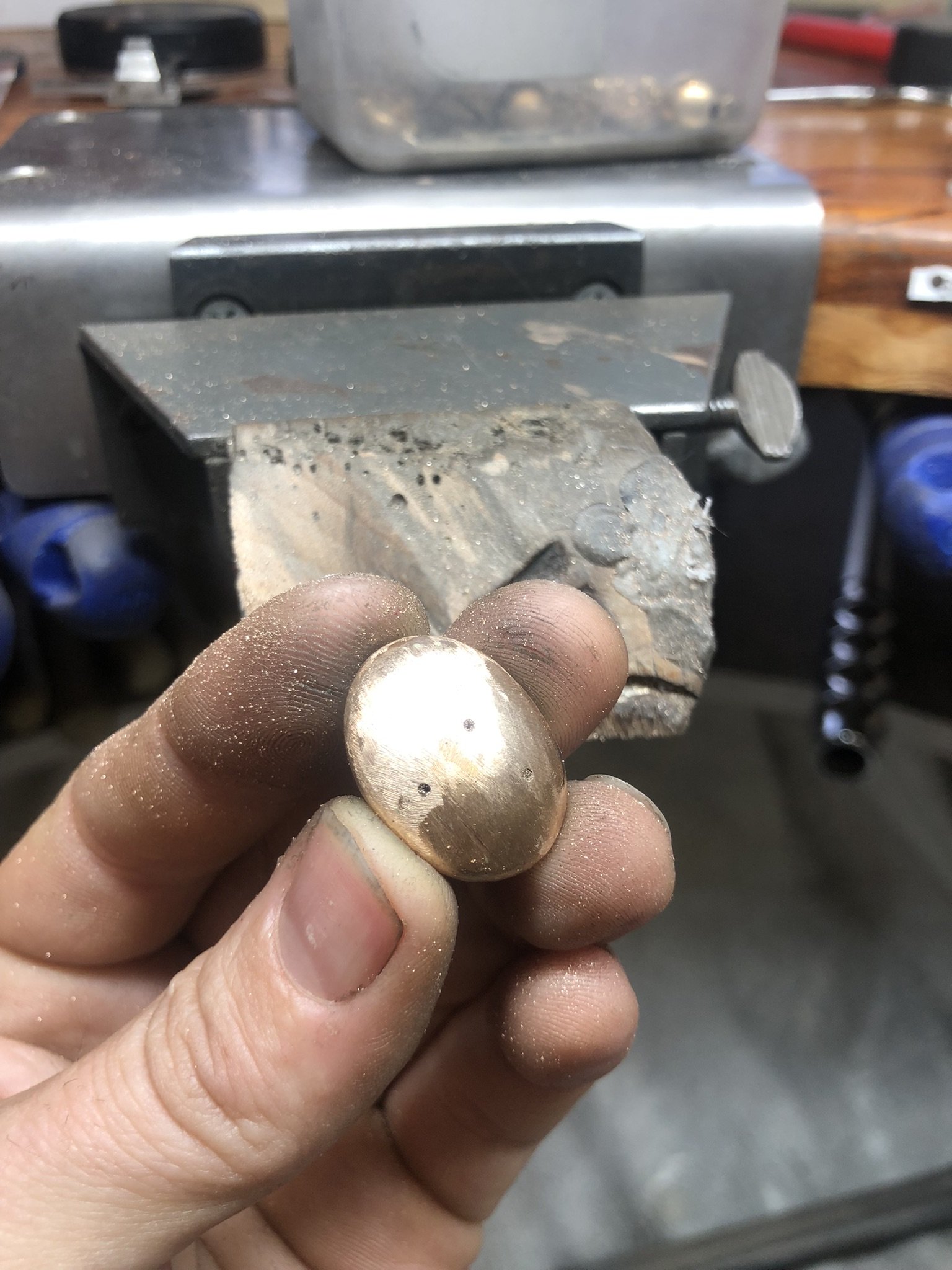 A person holding a small, round, shiny gold piece of jewellery in their fingers. The background shows a workbench with tools and equipment.