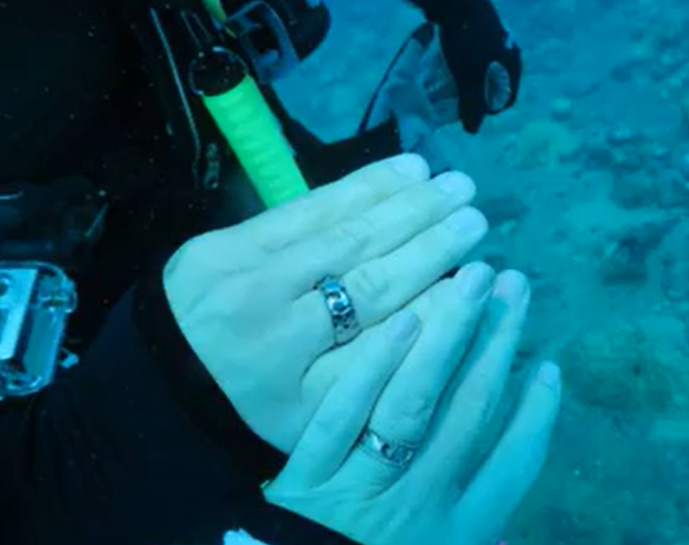 Close-up of a diver's hands underwater, with rings on fingers, and scuba gear visible in the background.