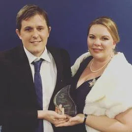 A man and woman dressed in formal attire holding an award together against a blue background.