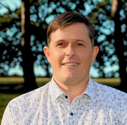 Portrait of a man with short brown hair, wearing a white patterned button-up shirt, outdoors with trees in the background.