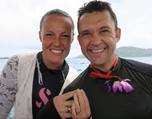 A smiling woman and man taking a selfie outdoors near water, with the woman dressed casually and the man wearing a black shirt with a red bow tie and purple flowers.