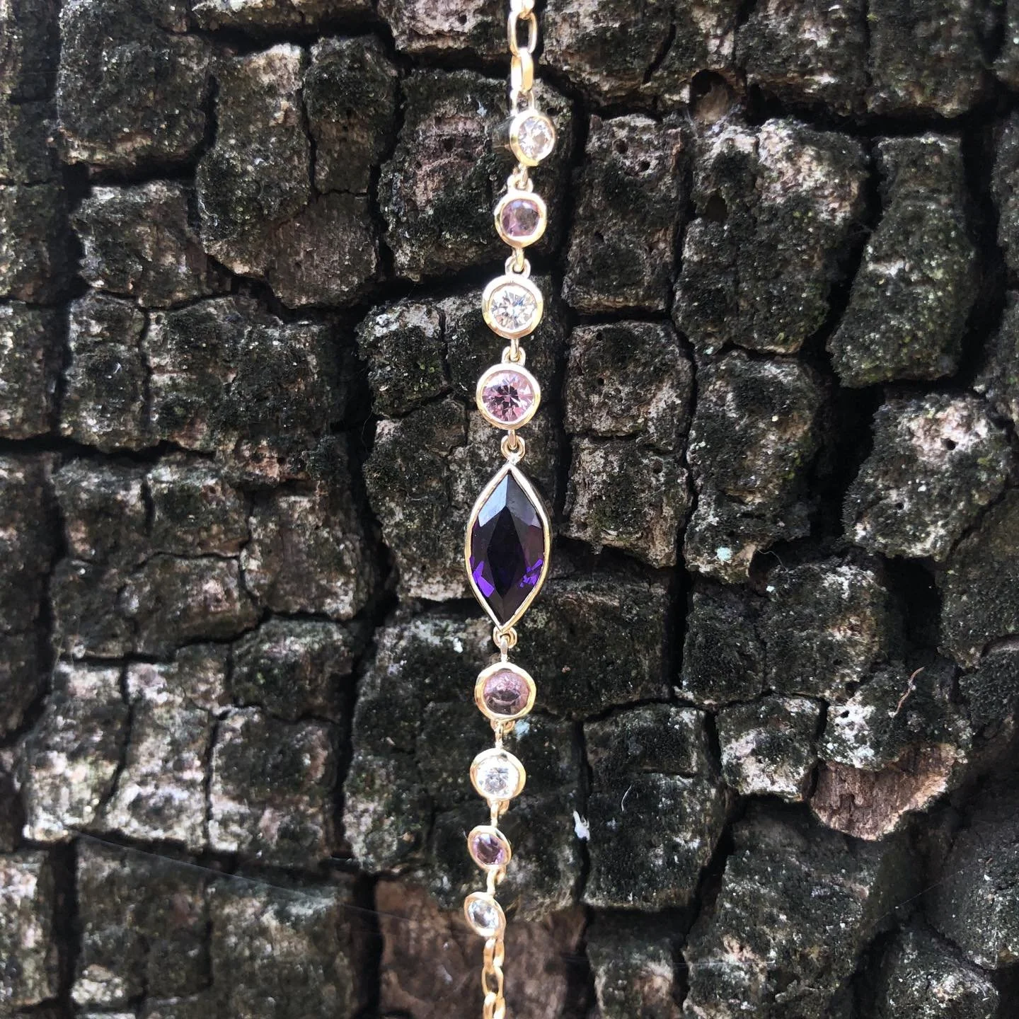 A delicate bracelet featuring round diamonds and one marquise-shaped purple amethyst stone, hanging against a rough, dark, volcanic rock background.