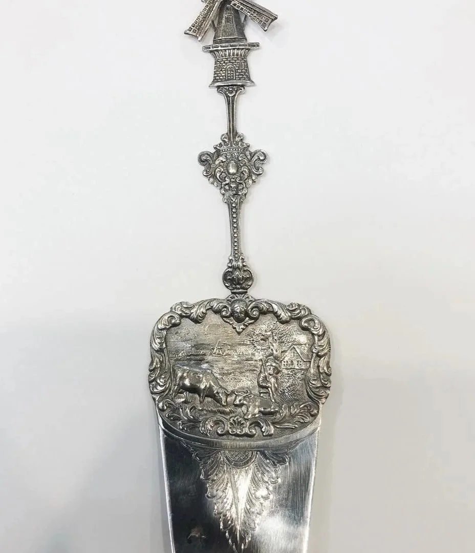 A decorative sterling silver cake server with an ornate handle and a detailed dutch scene.