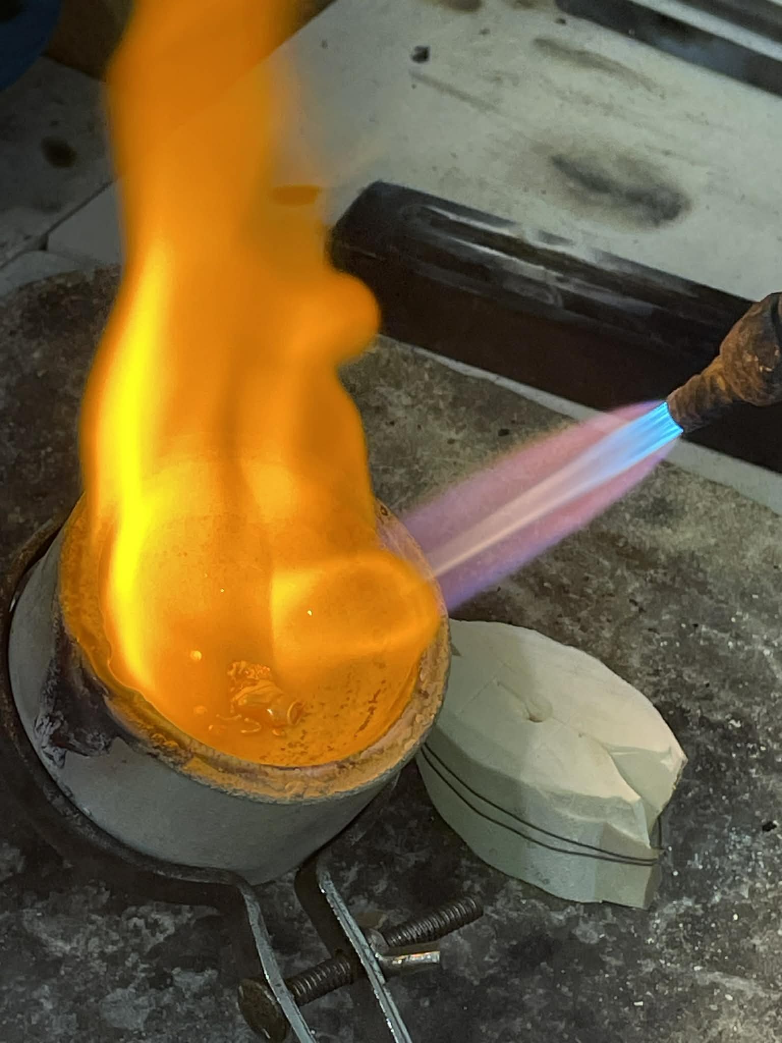 A person holding a blowtorch appears to be working on a hot glass object, possibly a glass sculpture or a piece of art, on a workbench. The scene is indoors with tools and materials visible in the background.