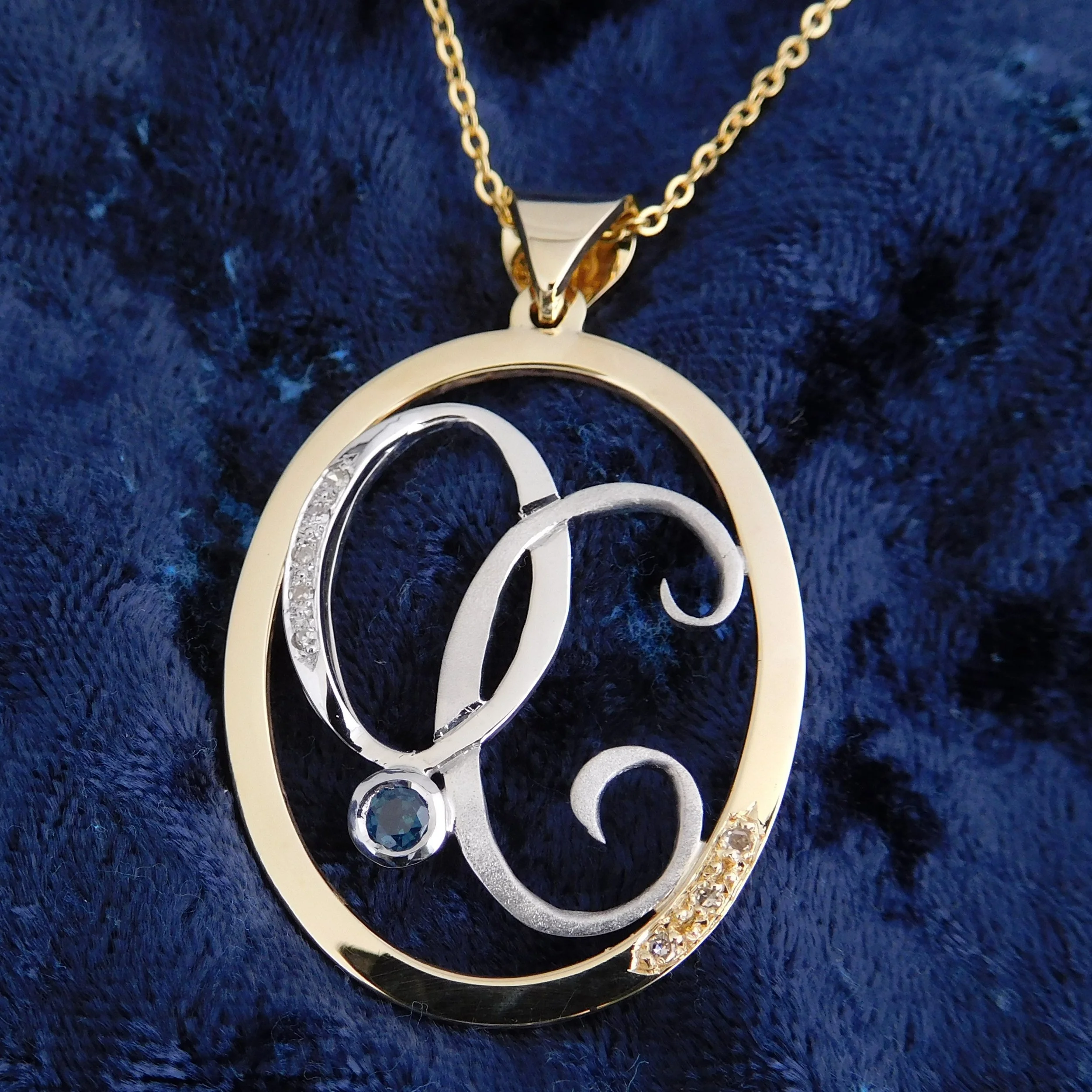 Finished two-tone gold monogram pendant with teal sapphire and diamonds on blue fabric