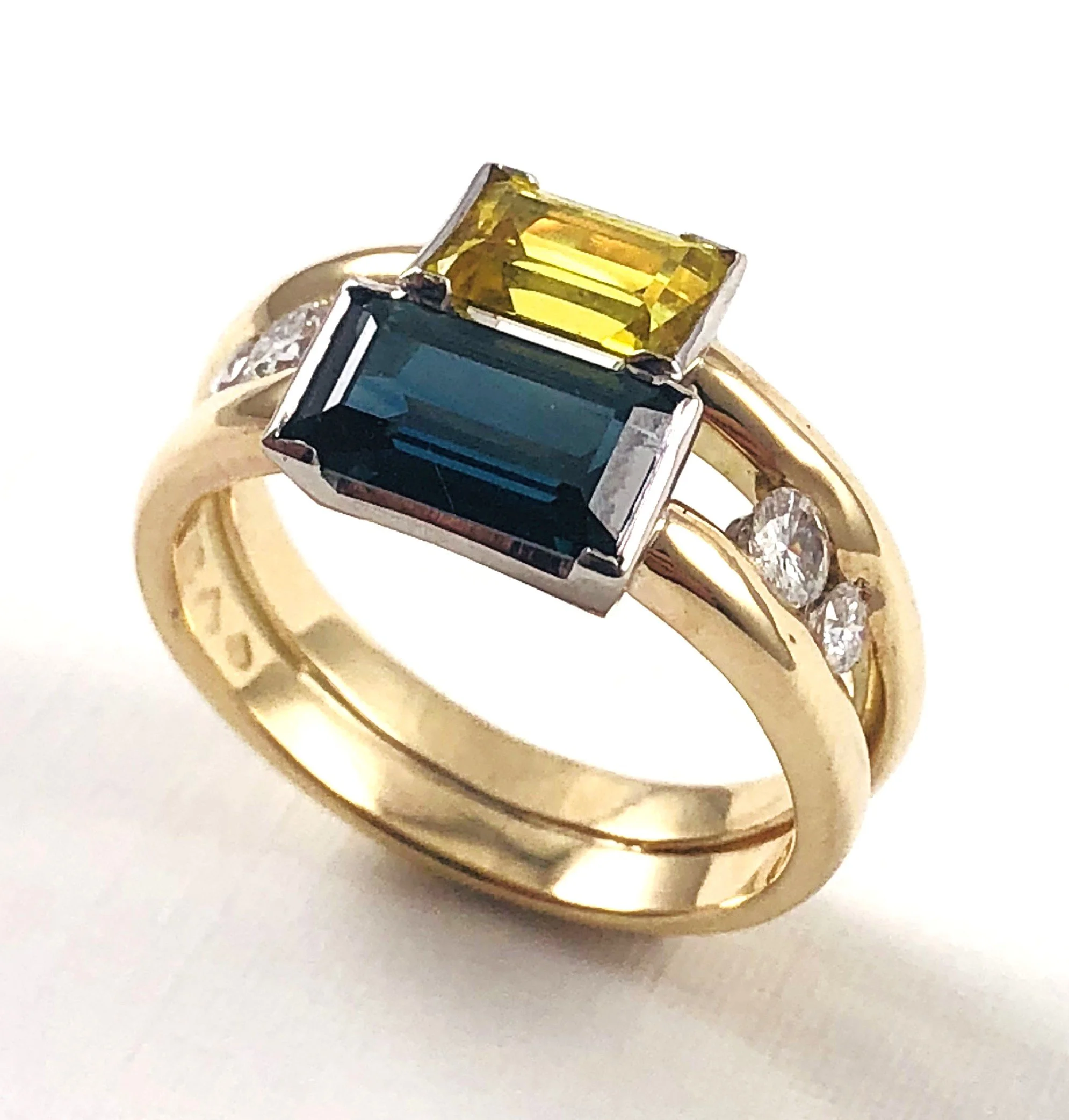 Close-up of rings, featuring a gold band with a large rectangular yellow gemstone and a black square stone set in a silver band, on a white surface.