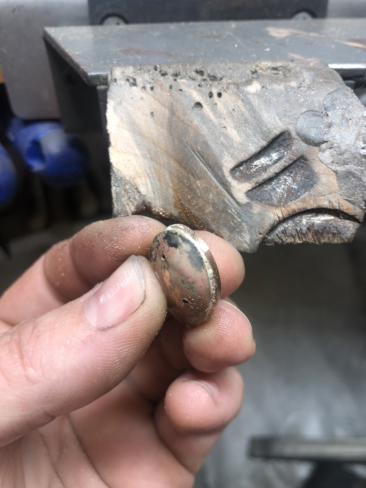 A person holding a small, round, metallic object in their fingers near a metal workpiece with grooves, in a workshop setting.