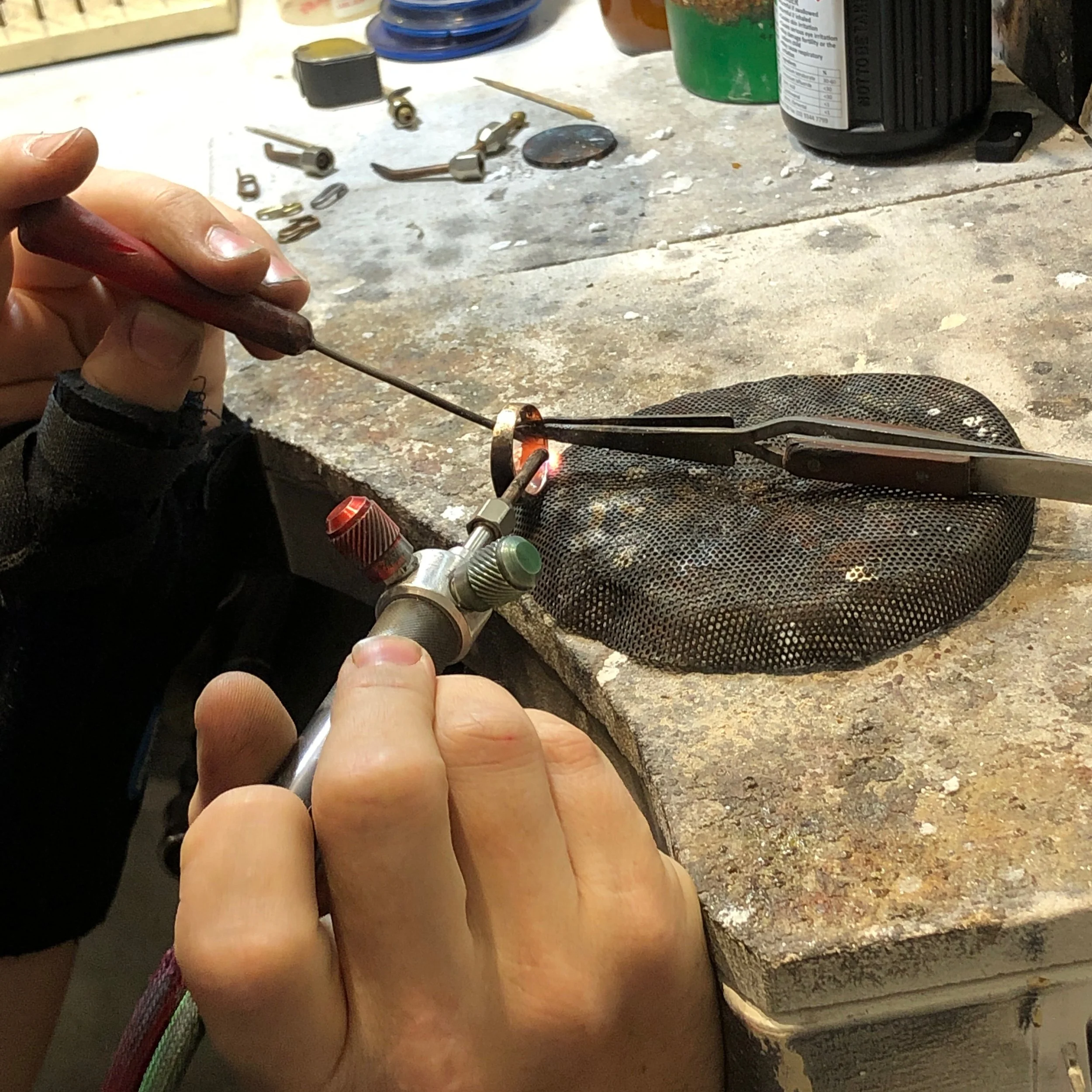 Close-up of a person holding a small tool or wire near a workbench with small metal parts, bottles, and containers scattered on the surface.