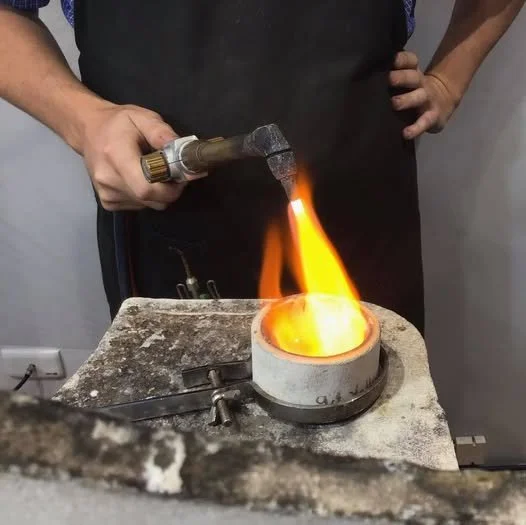 Jeweller using a blowtorch to heat and melt gold on a workbench.