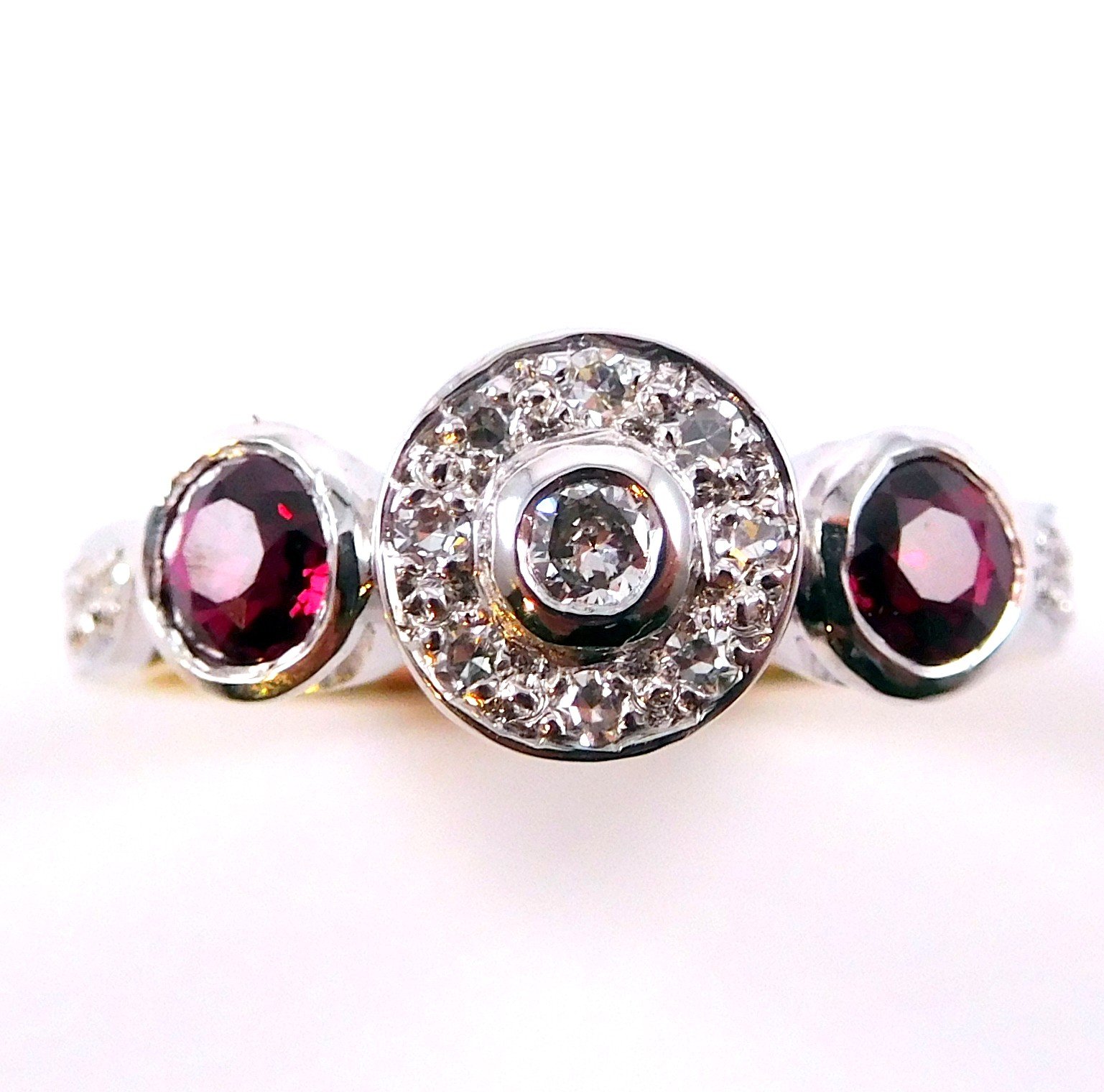 A ring with a large central clear gemstone surrounded by many small clear gemstones, flanked by two smaller red gemstones on either side.