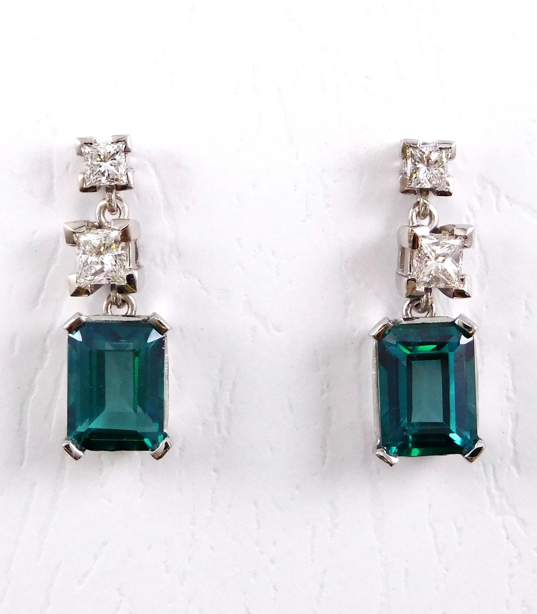 A pair of dangling earrings with large emerald-cut teal gemstones, supported by a metal setting with smaller square-cut diamonds above.