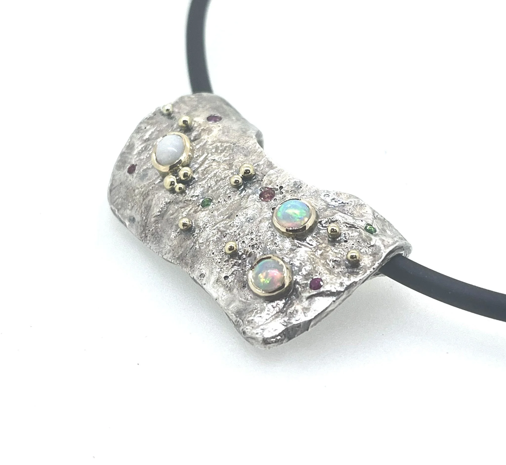 A silver arty pendant adorned with various small gemstones and a larger opals, connected to a black cord.