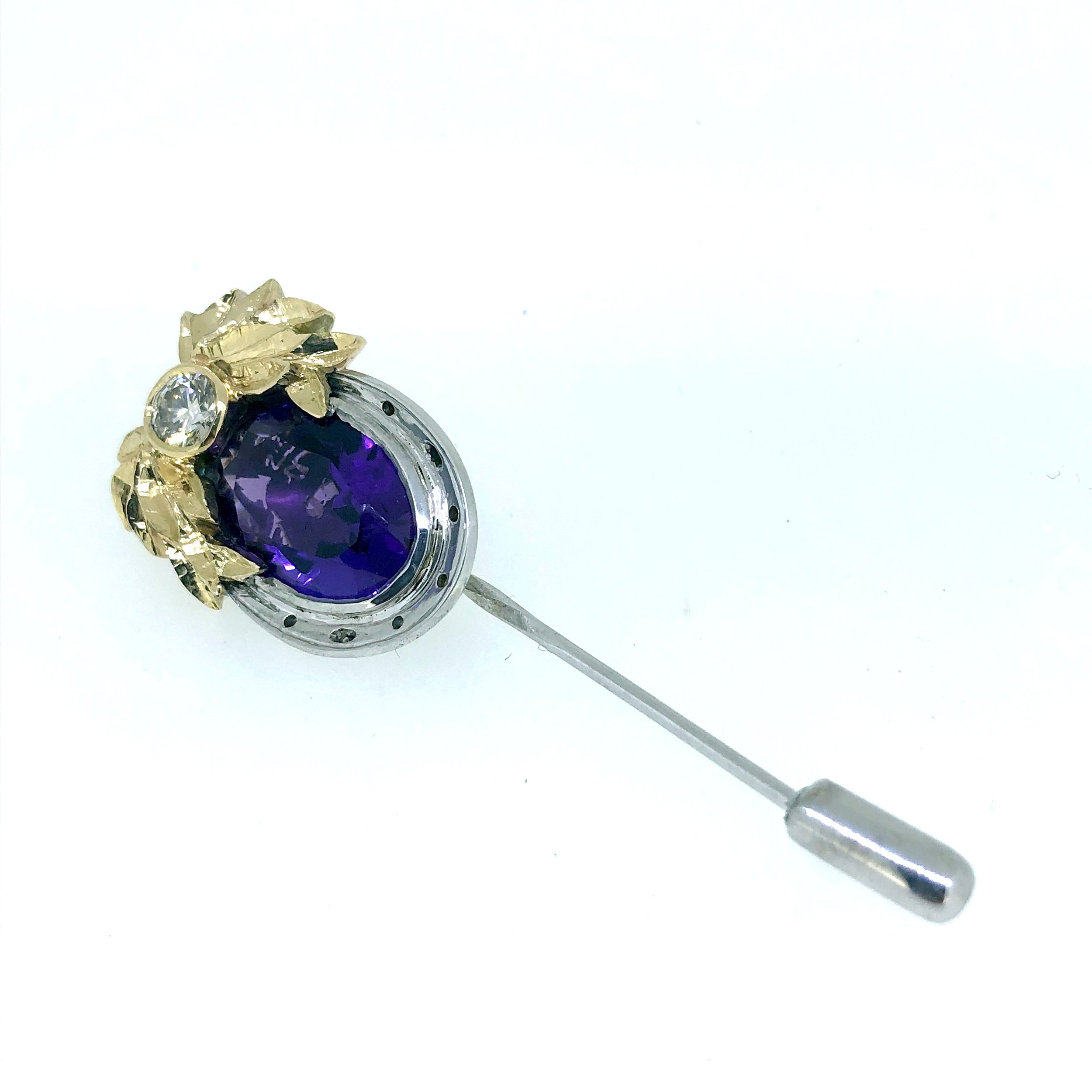 Close-up of a decorative silver hat pin, with a large purple gemstone, adorned with gold and clear gemstone accents,