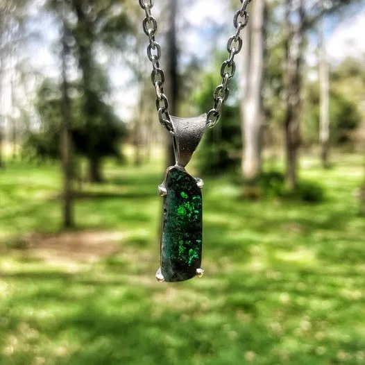 A necklace with a rectangular green and black gemstone pendant hanging from a chain, outdoors with trees and grass in the background