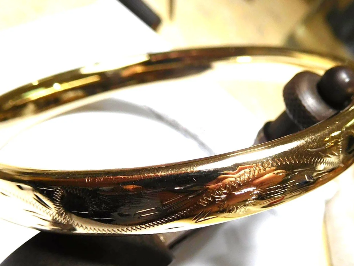 Close-up of a gold bangle with intricate engraved patterns, resting on a reflective surface.